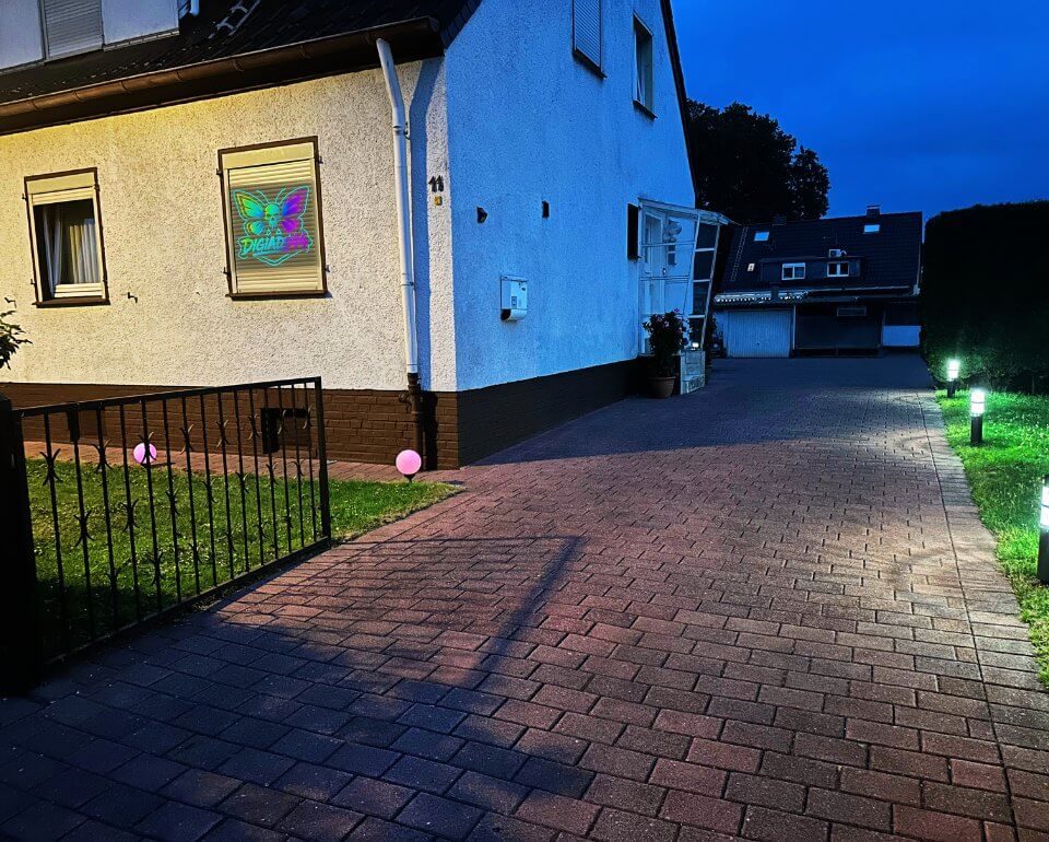 DigiAds44: Digitale Präsenz auch bei Nacht Abendansicht des Hauses DigiAds44 mit beleuchtetem Fensterlogo und gepflasterter Einfahrt, rosa Leuchte im Vorgarten