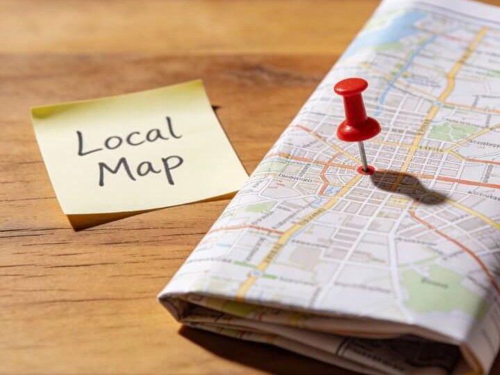 Gelbes "Local Map"-Post-it neben roter Stecknadel auf gefalteter Stadt-Karte auf Holztisch