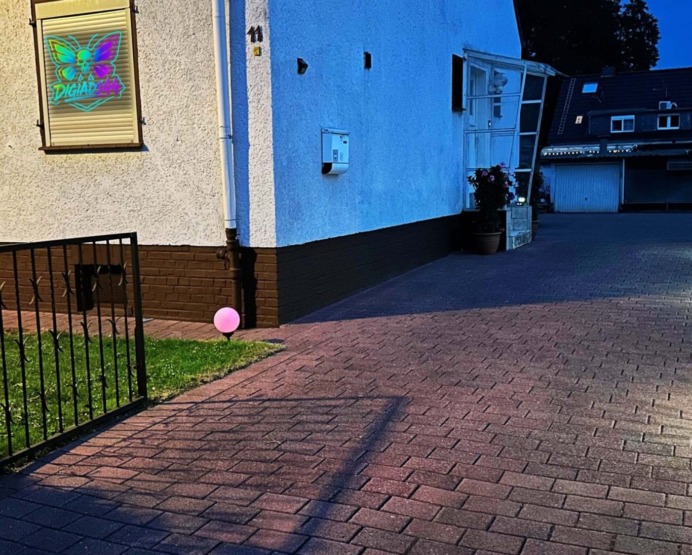 Abendansicht des Hauses DigiAds44 mit beleuchtetem Fensterlogo und gepflasterter Einfahrt, rosa Leuchte im Vorgarten